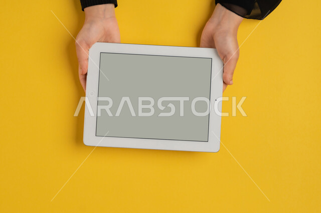 عرض شاشة رمادية فارغة، متابعة الدروس التعليمية، بورتريه علوي عامودي ليدين إمرأة عربية خليجية إماراتية تقوم بإنجاز المهام المطلوبة عن بعد، شابة سعودية ترتدي العباءة تستخدام جهاز التابليت، التعليم عن بعد بواسطة الانترنت، الاستمتاع بمشاهدة الافلام، خلفية صفراء