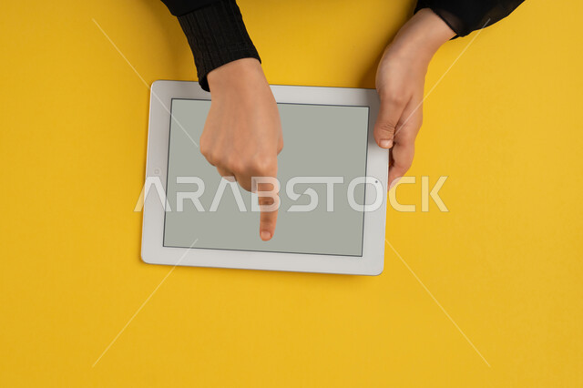 استخدام التكنولوجيا والتقنيات الحديثة والمتطورة، بورتريه علوي عامودي ليدين إمرأة عربية خليجية إماراتية تحمل التابليت، شابة سعودية ترتدي العباءة تقوم بتصفح مواقع التواصل الاجتماعي، القيام بعمليات التسويق الالكتروني، النقر على شاشة رمادية فارغة، خلفية صفراء