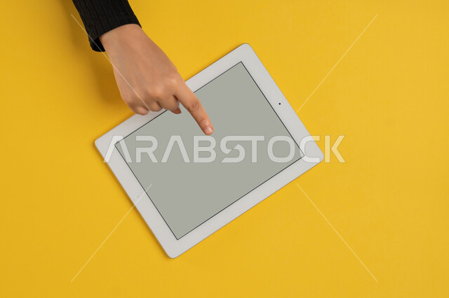 النقر على شاشة رمادية فارغة، دمج التكنولوجيا والتقنيات الحديثة والمتطورة بالعمل، بورتريه علوي عامودي ليدين إمرأة عربية خليجية إماراتية تحمل التابليت، شابة سعودية ترتدي العباءة تقوم بتصفح مواقع التواصل الاجتماعي، القيام بعمليات التسويق الالكتروني، خلفية صفراء