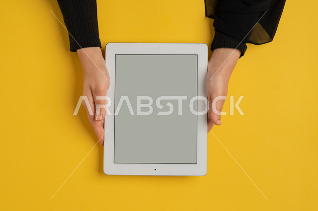 متابعة الدروس التعليمية أونلاين، بورتريه علوي عامودي ليدين إمرأة عربية خليجية إماراتية تقوم بإنجاز المهام المطلوبة عن بعد، شابة سعودية ترتدي العباءة تستخدام جهاز التابليت، التعليم عن بعد، الاستمتاع بمشاهدة الافلام، عرض شاشة رمادية فارغة، خلفية صفراء