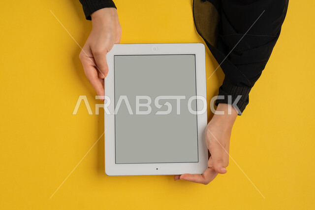 التعليم عن بعد بواسطة الانترنت، الاستمتاع بمشاهدة الافلام، عرض شاشة رمادية فارغة، متابعة الدروس التعليمية، بورتريه علوي عامودي ليدين إمرأة عربية خليجية إماراتية تقوم بإنجاز المهام المطلوبة عن بعد، شابة سعودية ترتدي العباءة تستخدام جهاز التابليت، خلفية صفراء
