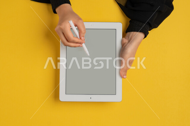 كتابة وتدوين الملاحظات باستخدام قلم التابليت الخاص، بورتريه علوي عامودي ليدين إمرأة عربية خليجية إماراتية تقوم بإنجاز المهام المطلوبة عن بعد، شابة سعودية ترتدي العباءة تستعمل جهاز الآيباد، رسم الصور والتصاميم الالكترونية، عرض شاشة رمادية فارغة، خلفية صفراء
