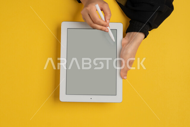 استخدام قلم التابلت لتدوين ملاحظات خاصة بالعمل، عرض شاشة رمادية فارغة، بورتريه علوي عامودي ليدين إمرأة عربية خليجية إماراتية تقوم بإنجاز المهام المطلوبة عن بعد، شابة سعودية ترتدي العباءة تستعمل جهاز الآيباد، رسم الصور والتصاميم الالكترونية، خلفية صفراء