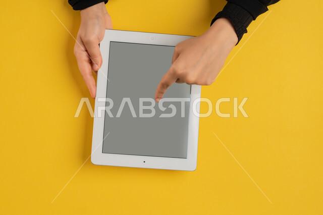 Clicking on a blank gray screen, integrating modern and advanced technology and techniques into work, vertical overhead portrait of the hands of an Arab Gulf Emirati woman holding a tablet, a young Saudi woman wearing an abaya browsing social media sites, doing electronic marketing operations, yellow background