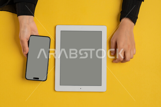 عرض شاشة رمادية فارغة، التسويق الالكتروني للتطبيقات، دمج التكنولوجيا الحديثة في الحياة اليومية والعملية، بورتريه علوي عامودي لايدي امرأة عربية خليجية سعودية تحمل الهاتف المحمول، شابة اماراتية تقوم باستخدام جهاز التابلت، خلفية صفراء