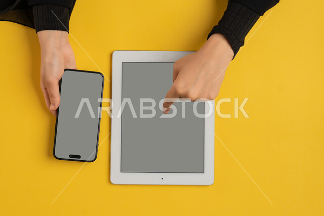 Integrating modern technology into daily and business life, blank gray screen display, e-marketing for applications, vertical overhead portrait of a Saudi Arabian Gulf woman's hands tapping on a tablet, young Emirati woman using a mobile phone, yellow background