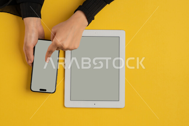 E-marketing for applications, blank gray screen display, integration of modern technology into daily and business life, vertical overhead portrait of a Saudi Arabian Gulf woman's hands tapping on a mobile phone, young Emirati woman using a tablet, yellow background