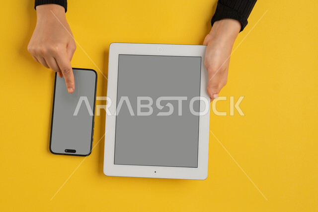 التسويق الالكتروني للتطبيقات، عرض شاشة رمادية فارغة، دمج التكنولوجيا الحديثة في الحياة اليومية والعملية، بورتريه علوي عامودي لايدي امرأة عربية خليجية سعودية تقوم بالنقرعلى الهاتف المحمول، شابة اماراتية تستخدام جهاز التابلت، خلفية صفراء