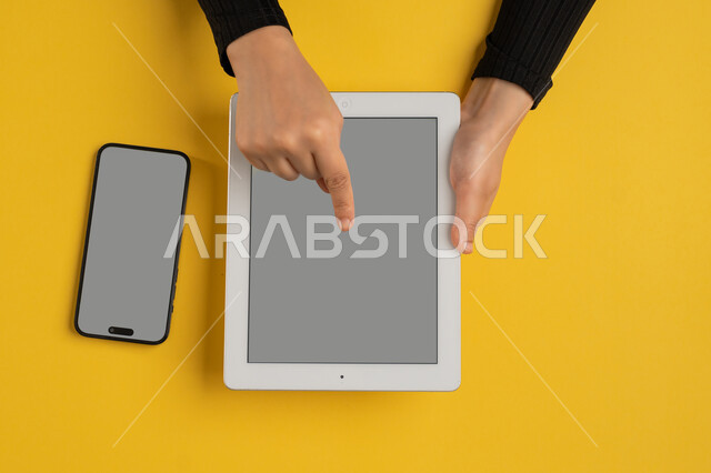 Integrating modern technology into daily and business life, blank gray screen display, e-marketing for applications, vertical overhead portrait of a Saudi Arabian Gulf woman's hands tapping on a tablet, young Emirati woman using a mobile phone, yellow background