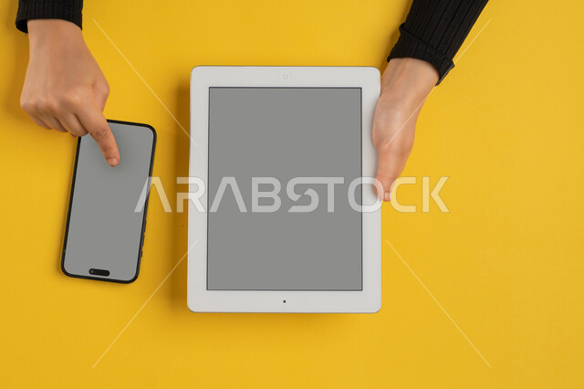 E-marketing for applications, blank gray screen display, integration of modern technology into daily and business life, vertical overhead portrait of a Saudi Arabian Gulf woman's hands tapping on a mobile phone screen, young Emirati woman using a tablet, yellow background