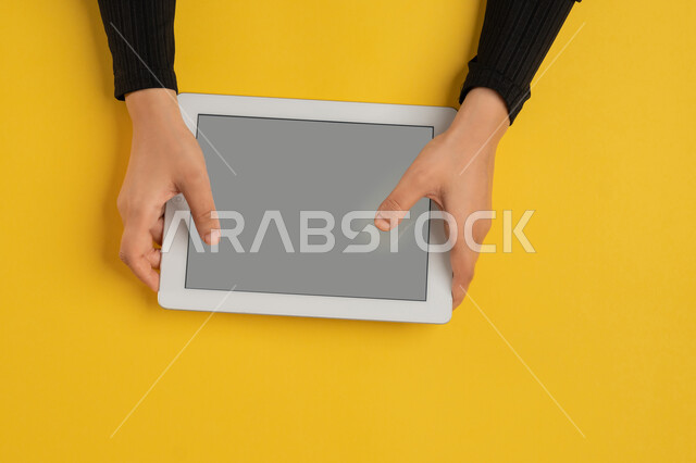 Using modern technology, following educational lessons, enjoying watching movies, vertical overhead portrait of a Saudi Arabian Gulf woman's hands tapping on a tablet, young Emirati woman playing video games, blank gray screen display, yellow background