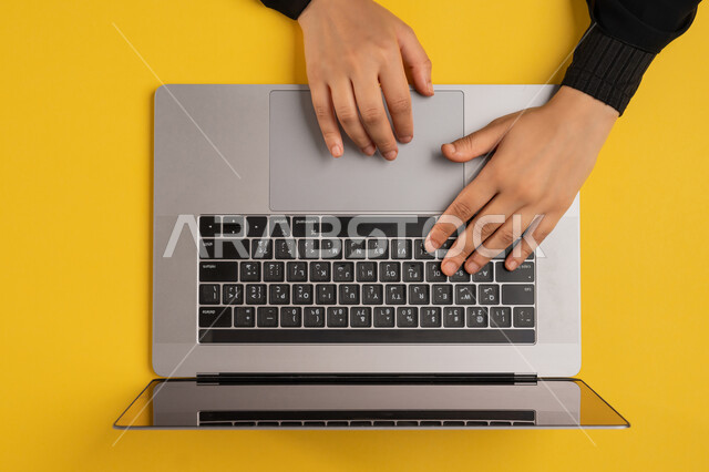 Accomplishing required tasks remotely, integrating modern technology into work life, vertical overhead portrait of a Gulf Arab Emirati woman working on laptop, young Saudi woman wearing abaya typing on keyboard, concept of business management and achieving goals, yellow background