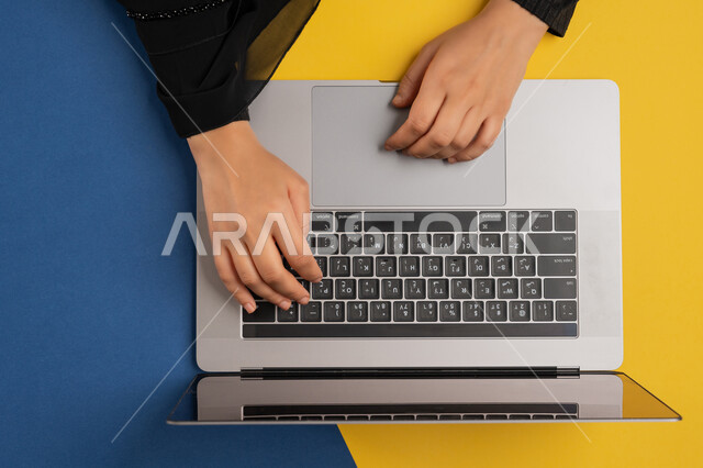 Completing required tasks remotely, vertical overhead portrait of a Gulf Arab Emirati woman working on laptop, young Saudi woman wearing abaya typing on keyboard, integrating modern technology into business life, concept of business management and achieving goals, colorful background