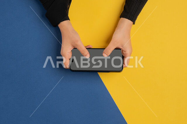 القيام بعمليات التسويق الالكتروني، النقر على شاشة رمادية فارغة، دمج التكنولوجيا الحديثة المتطورة بالحياة اليومية والعملية، بورتريه علوي عامودي لايدي امرأة عربية خليجية إماراتية تحمل الهاتف المحمول، شابة سعودية تقوم بتصفح مواقع التواصل الاجتماعي، خلفية ملونة