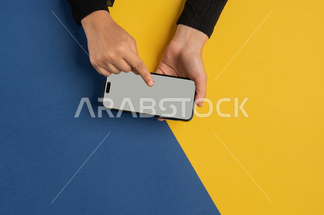 القيام بعمليات التسويق الالكتروني، النقر على شاشة رمادية فارغة، دمج التكنولوجيا الحديثة المتطورة بالحياة اليومية والعملية، بورتريه علوي عامودي لايدي امرأة عربية خليجية إماراتية تحمل الهاتف المحمول، شابة سعودية تقوم بتصفح مواقع التواصل الاجتماعي، خلفية ملونة