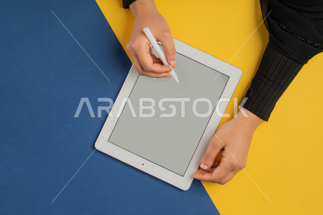 رسم الصور والتصاميم الالكترونية، عرض شاشة رمادية فارغة، استخدام قلم التابلت لتدوين ملاحظات خاصة بالعمل، بورتريه علوي عامودي لايدي إمرأة عربية خليجية إماراتية تقوم بإنجاز المهام المطلوبة عن بعد، شابة سعودية ترتدي العباءة تستعمل جهاز الآيباد، خلفية ملونة