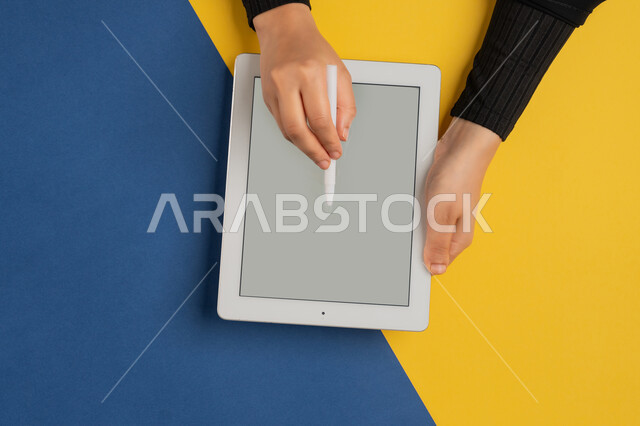 Blank gray screen display, using tablet pen to take notes for work, vertical overhead portrait of Emirati Gulf Arab woman hands doing tasks remotely, young Saudi woman wearing abaya using iPad, drawing pictures and electronic designs, colorful background