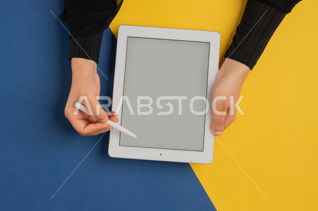 استخدام قلم التابلت لتدوين ملاحظات خاصة بالعمل، عرض شاشة رمادية فارغة، بورتريه علوي عامودي لايدي إمرأة عربية خليجية إماراتية تقوم بإنجاز المهام المطلوبة عن بعد، شابة سعودية ترتدي العباءة تستعمل جهاز الآيباد، رسم الصور والتصاميم الالكترونية، خلفية ملونة