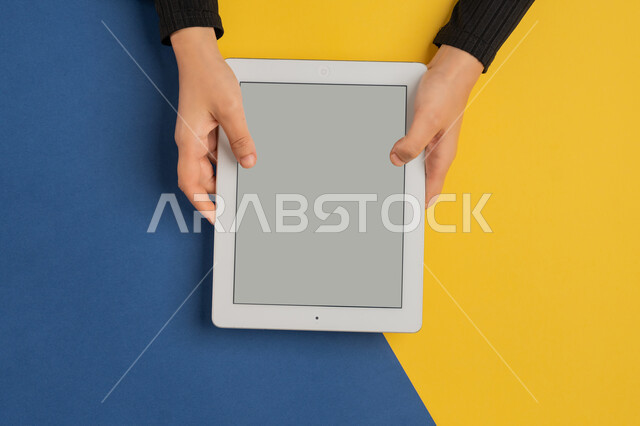 Using modern technology, following educational lessons, enjoying watching movies, vertical overhead portrait of a Saudi Arabian Gulf woman's hands tapping on a tablet, young Emirati woman playing video games, blank gray screen display, colorful background