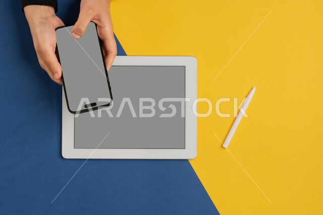 Integrating modern technology into daily and business life, blank gray screen display, e-marketing for applications, vertical overhead portrait of a Saudi Arabian Gulf woman's hands tapping on a mobile phone screen, young Emirati woman using a tablet, colorful background