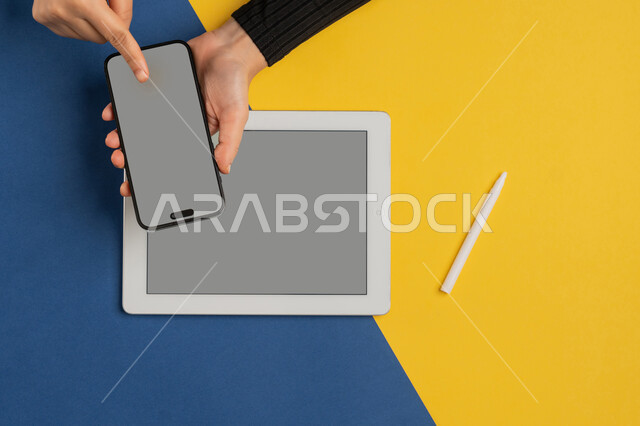 Blank gray screen display, integration of modern technology into daily and business life, e-marketing for applications, vertical overhead portrait of a Saudi Arabian Gulf woman's hands tapping on a mobile phone screen, young Emirati woman using a tablet, colorful background