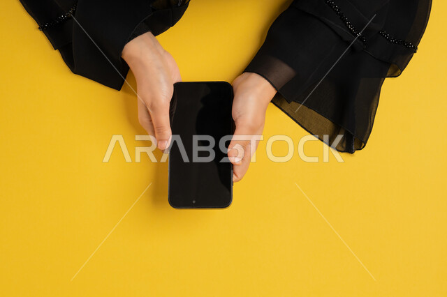 دمج التكنولوجيا الحديثة في الحياة اليومية والعملية، التسويق الالكتروني للتطبيقات، عرض شاشة سوداء فارغة، بورتريه علوي عامودي لايدي امرأة عربية خليجية سعودية تقوم بتقديم خدمات الموبايل، شابة اماراتية تستخدام الهاتف المحمول، خلفية صفراء