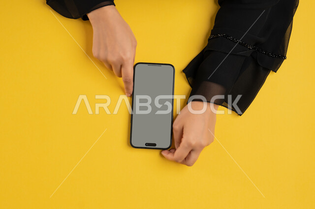 التسويق الالكتروني للتطبيقات، عرض شاشة رمادية فارغة، دمج التكنولوجيا الحديثة في الحياة اليومية والعملية، بورتريه علوي عامودي لايدي امرأة عربية خليجية سعودية تقوم بتقديم خدمات الموبايل، شابة اماراتية تستخدام الهاتف المحمول، خلفية صفراء