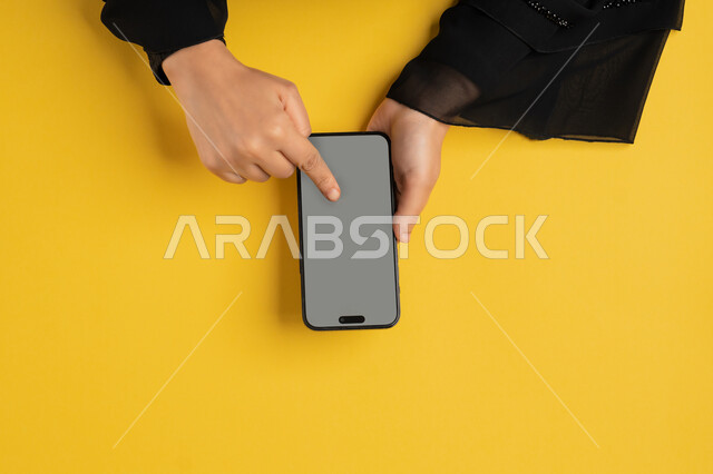 القيام بعمليات التسويق الالكتروني، النقر على شاشة رمادية فارغة، دمج التكنولوجيا الحديثة المتطورة بالحياة اليومية والعملية، بورتريه علوي عامودي لايدي امرأة عربية خليجية إماراتية تستخدم الهاتف المحمول، شابة سعودية تقوم بتصفح مواقع التواصل الاجتماعي، خلفية صفراء