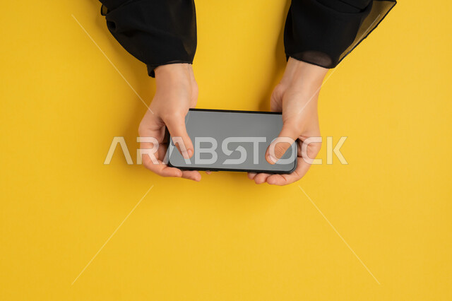 النقر على شاشة رمادية فارغة، القيام بعمليات التسويق الالكتروني، دمج التكنولوجيا الحديثة المتطورة بالحياة اليومية والعملية، بورتريه علوي عامودي لايدي امرأة عربية خليجية إماراتية تستخدم الهاتف المحمول، شابة سعودية تقوم بتصفح مواقع التواصل الاجتماعي، خلفية صفراء