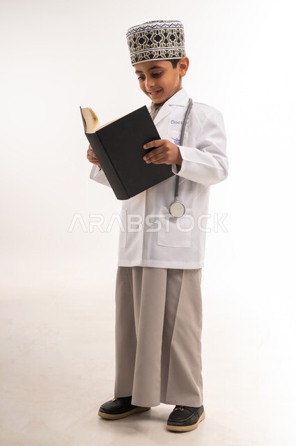 التحضير للحصول على مهنة الطب، مفهوم التثقف والتعلم الذاتي، اختيار وتحديد المهنة المستقبلية منذ الصغر، بورتريه لولد عربي خليجي عماني مبتسم يرتدي الكمة والمعطف الأبيض يضع السماعات الطبية يقوم بقراءة كتاب، الطول الكامل للجسم، خلفية بيضاء