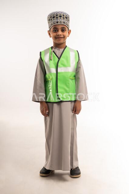 مفهوم العمل في القطاع الهندسي، اختيار وتحديد المستقبل منذ الصغر، ايماءات الشغف لتحقيق حلم الطفولة، بورتريه لولد عربي خليجي عماني مبتسم يرتدي الكمه وسترة الحماية يقف بإستقامة وينظر إلى الكاميرا بتعابير السعادة والسرور، الطول الكامل للجسم، خلفية بيضاء