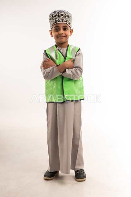 تحديد واختيار المستقبل منذ الصغر، مفهوم العمل في القطاع الهندسي، ايماءات الشغف لتحقيق حلم الطفولة، بورتريه لولد عربي خليجي عماني مبتسم يرتدي الكمه وسترة الحماية يقف بإستقامة وايدي متقاطعة وينظر إلى الكاميرا بتعابير السعادة، الطول الكامل للجسم، خلفية بيضاء