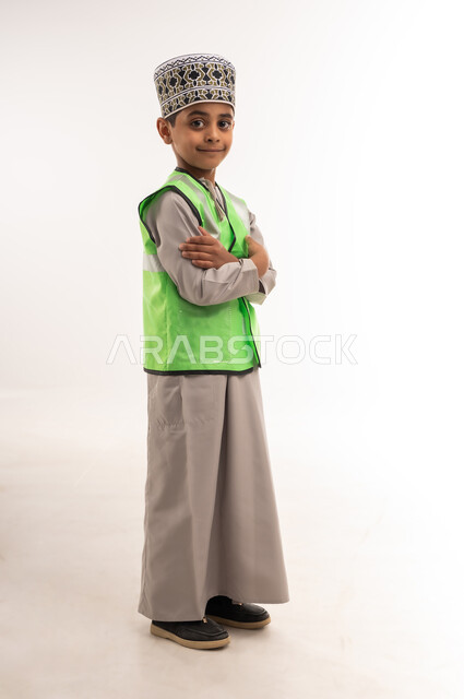 اختيار وتحديد المستقبل منذ الصغر، مفهوم العمل في القطاع الهندسي، ايماءات الشغف لتحقيق حلم الطفولة، بورتريه لولد عربي خليجي عماني مبتسم يرتدي الكمه وسترة الحماية يقف بإستقامة وايدي متقاطعة وينظر إلى الكاميرا بتعابير السعادة، الطول الكامل للجسم، خلفية بيضاء