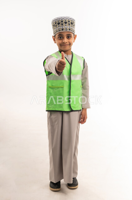 Gestures of passion to achieve childhood dream, defining and choosing the future since childhood, concept of working in the engineering sector, portrait of a smiling Arab Gulf Omani boy wearing a mask and a protective vest standing straight and raising his thumb with expressions of satisfaction and admiration, full body length, white background
