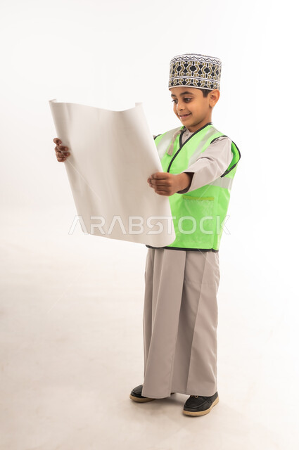 Follow-up and auditing of engineering projects and plans, the concept of work in the engineering sector, choosing and determining the future profession from a young age, portrait of a smiling Gulf Arab Omani boy wearing a sleeve and a protective vest holding a plan in his hand, full body length, white background