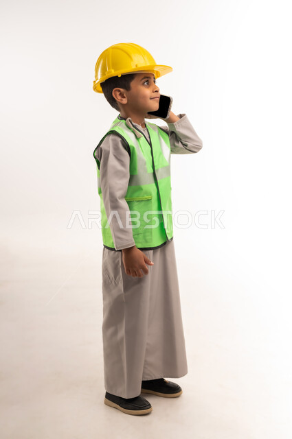 Dreaming of working in the engineering sector, supervising engineering projects and construction works, choosing and determining the future profession since childhood, portrait of a smiling Gulf Arab Omani boy wearing a helmet and a protective vest making a call on a mobile phone, full body length, white background