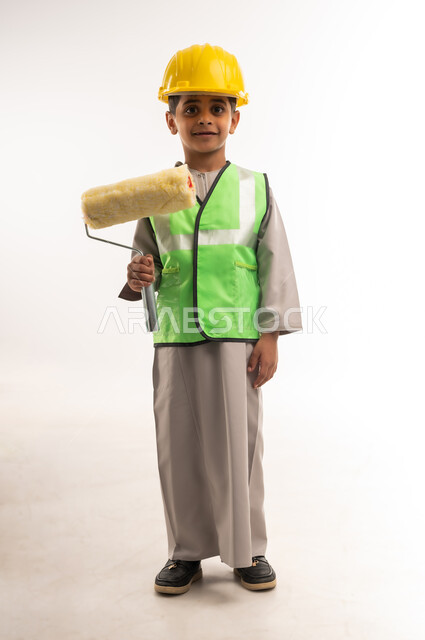 مفهوم العمل في القطاع الهندسي، أعمال الترميم وتشطيبات البناء، اختيار وتحديد المستقبل منذ الصغر، ايماءات الشغف لتحقيق حلم الطفولة، بورتريه لولد عربي خليجي عماني يرتدي الستره وخوذة الحماية يحمل بيده بكرة الدهان، الطول الكامل للجسم، خلفية بيضاء