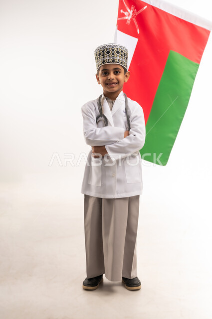 Love of the homeland and belonging to it, choosing and determining the future profession from a young age, portrait of a smiling Gulf Arab Omani boy wearing a turban and a white coat, wearing stethoscopes, standing with crossed hands and behind him the flag of the Sultanate of Oman, gestures of pride and honor, full body length, white background