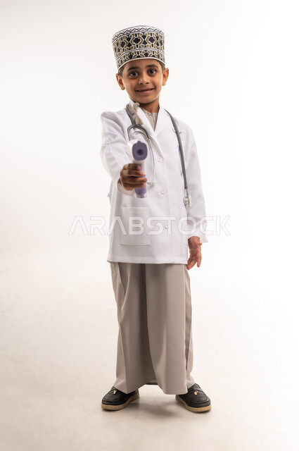 الإلتزام بإجراءات الوقاية من فايروس كورونا، بورتريه لولد عربي خليجي عماني يرتدي الكمة والمعطف الأبيض يضع سماعات الطبيب ويحمل بيده ميزان الحرارة الرقمي، مفهوم الطب والرعاية الصحية، اختيار وتحديد المهنة المستقبلية، الطول الكامل للجسم، خلفية بيضاء