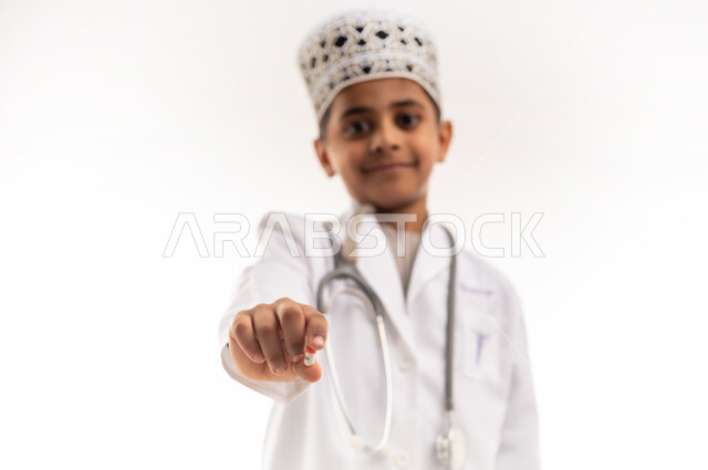 Providing health care and services, gestures of passion to achieve childhood dream and work in clinics and hospitals, close-up portrait of a smiling Gulf Arab Omani boy wearing a mask and white coat, wearing stethoscopes, holding a medicine capsule in his hand, choosing and determining the future profession since childhood, white background