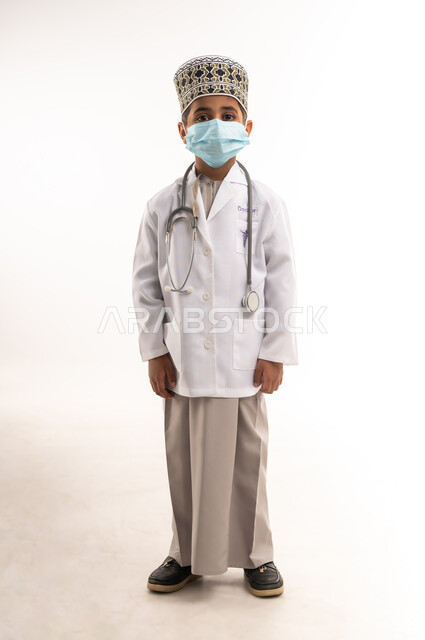 الاحترازات الوقائية ضد كوفيد 19، مفهوم الطب والرعاية الصحية، طموحات وأهداف مستقبلية، بورتريه لولد عربي خليجي عماني يرتدي يرتدي الكمة والمعطف الأبيض ويضع الكمامة وسماعات الطبيب، اختيار وتحديد مهنة المستقبل منذ الصغر، الطول الكامل للجسم، خلفية بيضاء