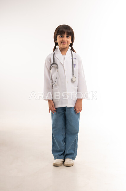 Hopes, ambitions and goals for the future, striving and working hard to achieve a childhood dream, choosing a future profession from a young age, portrait of a smiling Gulf Arab Omani girl wearing a white coat and a stethoscope, a Saudi girl standing straight and looking at the camera, full body length, white background