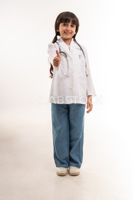 الاجتهاد والسعي لتحقيق حلم الطفولة، أهداف و طموحات للمستقبل، اختيار المهنه المستقبلية منذ الصغر، بورتريه لفتاة عربية خليجية عمانية ترتدي المعطف الأبيض وسماعة الطبيب، بنت سعودية ترفع اصبع الإبهام بايماءات الرضا والإعجاب، الطول الكامل للجسم، خلفية بيضاء