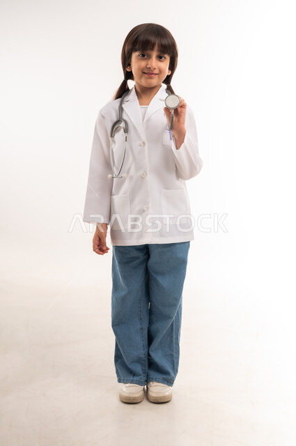 أهداف و طموحات للمستقبل، اختيار المهنه المستقبلية منذ الصغر، قياس نبضات القلب باستخدام السماعات الطبية، الاجتهاد والسعي لتحقيق حلم الطفولة، بورتريه لفتاة عربية خليجية عمانية ترتدي المعطف الأبيض، بنت سعودية تقف باستقامة، الطول الكامل للجسم، خلفية بيضاء