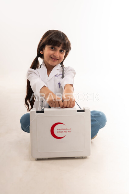 Pursuing a medical career, helping patients in emergency situations, future ambitions to work in hospitals, portrait of an Arab Gulf Omani girl wearing a coat and a stethoscope, a Saudi girl sitting and looking at the camera with gestures of happiness and next to her a first aid kit, white background