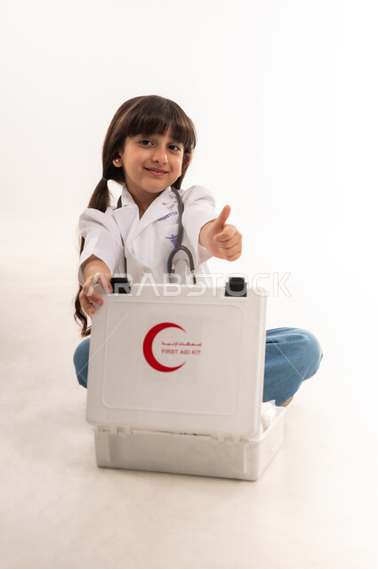 رفع اصبع الإبهام بايماءات الرضا و الإعجاب، مساعدة المرضى في الحالات الطارئة، الطموحات المستقبلية بالعمل في المستشفيات، بورتريه لفتاة عربية خليجية عمانية ترتدي المعطف وسماعة الطبيب، بنت سعودية تجلس وتقوم بفتح حقيبة الاسعافات الاولية، خلفية بيضاء