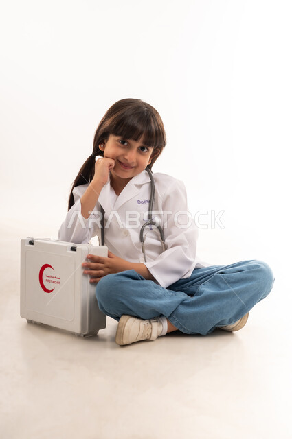 Dreaming of a medical career, helping patients in emergency situations, future ambitions to work in hospitals, portrait of an Arab Gulf Omani girl wearing a coat and a stethoscope, a Saudi girl sitting and looking at the camera with gestures of happiness and next to her is a first aid kit, white background