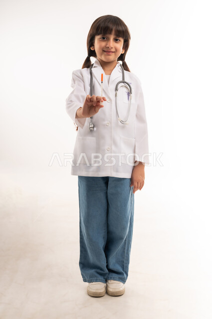 السعي للحصول على الشهادات العليا، أمنيات و آمال مستقبيله بالعمل في المختبرات والتحاليل، مفهوم الطب والرعاية الصحية، بورتريه لفتاة عربية خليجية عمانية مبتسمة ترتدي المعطف والسماعات الطبية، بنت سعودية تحمل بيدها حقنة طبية، الطول الكامل للجسم، خلفية بيضاء