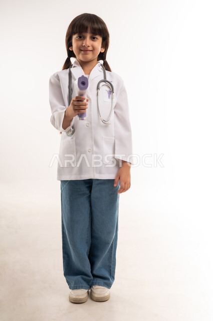 الالتزام بإجراءات السلامة والوقاية من فايروس كورونا، ، بورتريه لفتاة عربية خليجية عمانية ترتدي المعطف والسماعات الطبية تحمل بيدها ميزان الحرارة الرقمي، بنت سعودية تنظر إلى الكاميرا وتقف باستقامة، الطموح بالعمل في المشافي، الطول الكامل للجسم، خلفية بيضاء