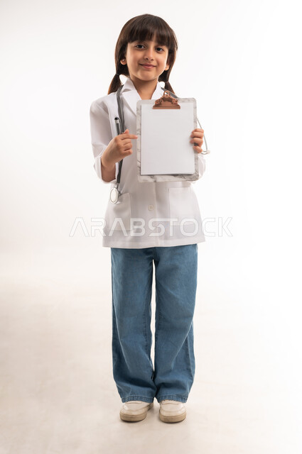 Preparing medical reports and reviewing patient records, determining future career from childhood, future goals of working in hospitals, portrait of a smiling Omani Gulf Arab girl wearing a white coat and a stethoscope, Saudi girl displaying an empty medical record, full body length, white background